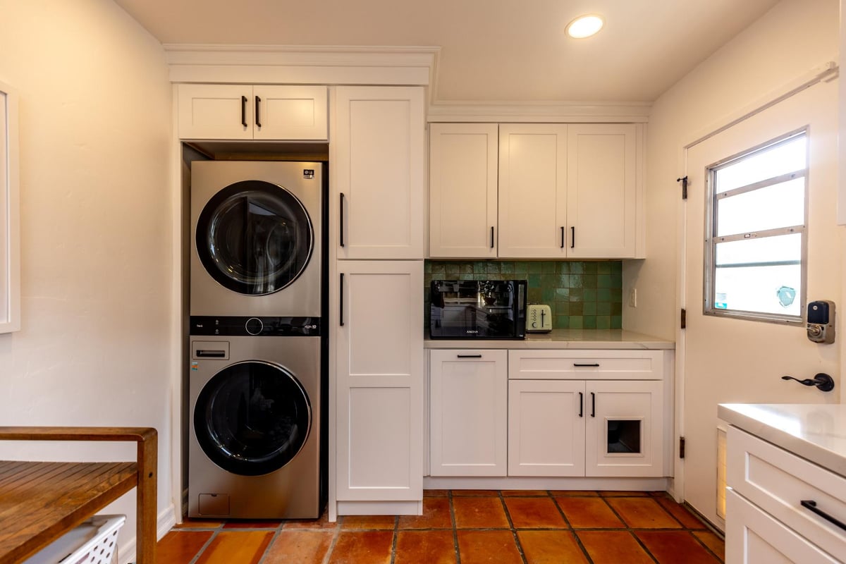 A laundry area within a kitchen, with white cabinets, a stacked washer and dryer set, a green tile backsplash, and terracotta floor tiles.